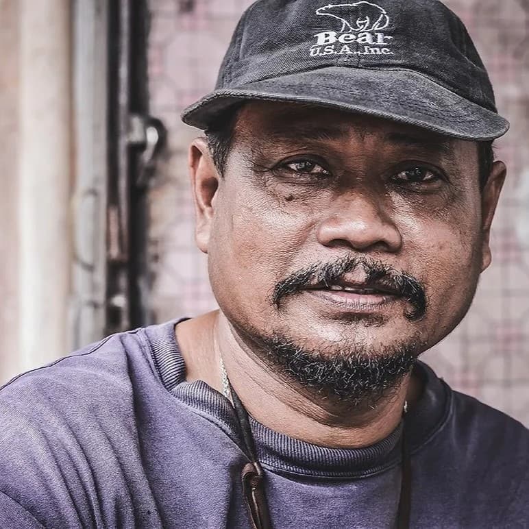 Guy with hat and necklace looking at camera.