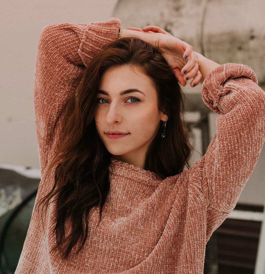Girl with black hair smiling at camera with both hand behind head.