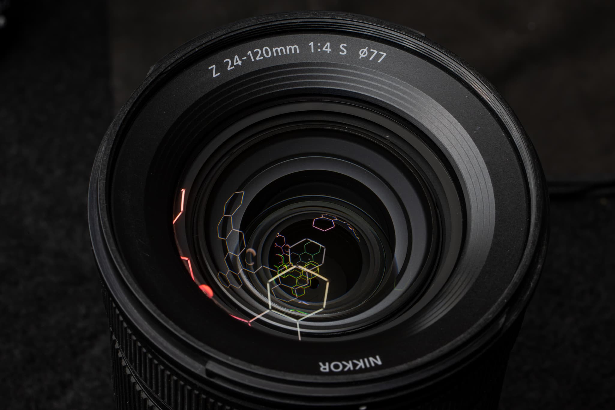 Tight accent shot of the expansive 77mm front glass element of the Nikon Z 24-120mm f/4 S catching a harsh, angled grid light to highlight the deep greenish and reddish reflections of the advanced Nano Crystal Coat and ARNEO Coat