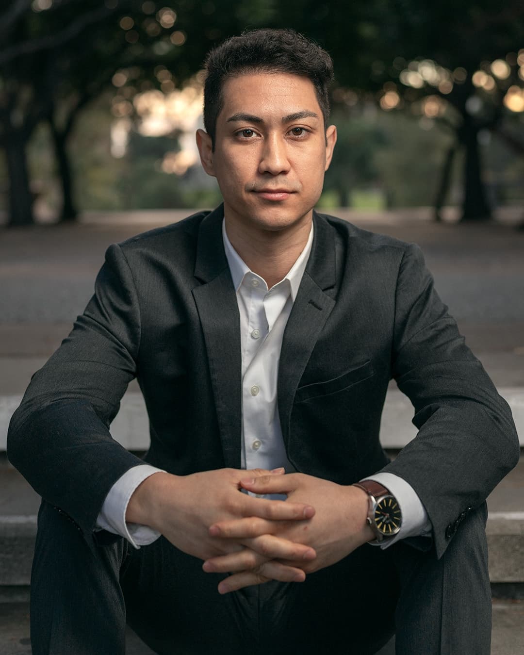Man posing in suit sitting hands grasped.